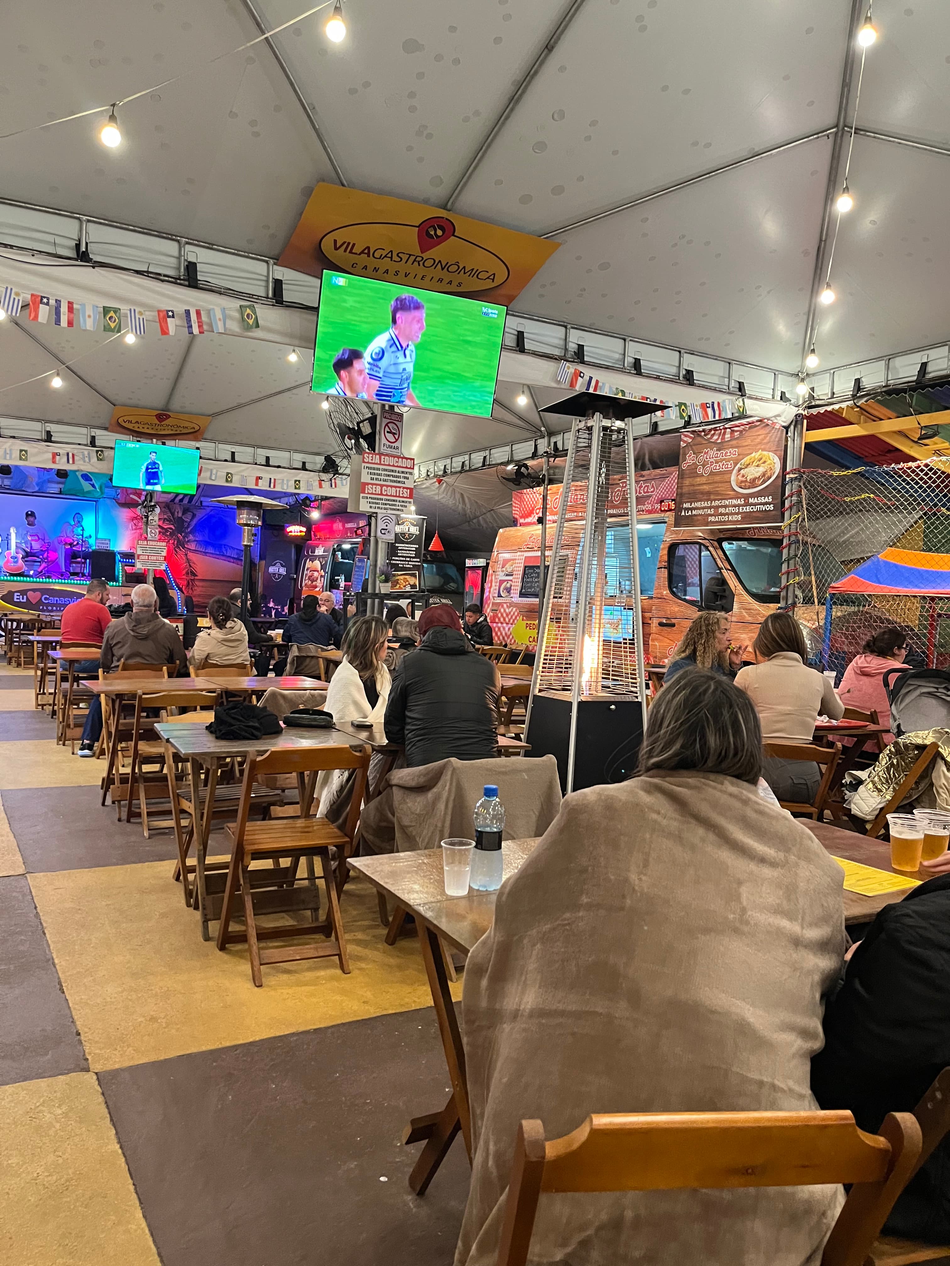 Ambiente interno da Vila Gastronômica com mesas de madeira e food trucks